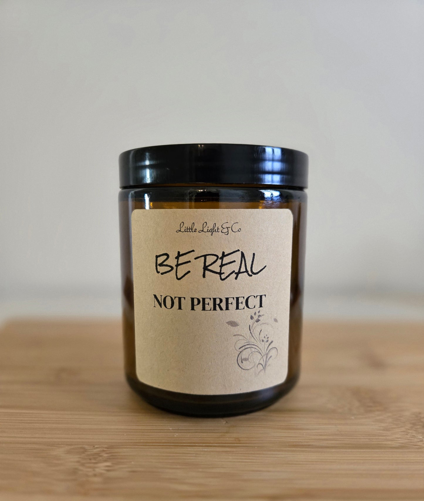 Candle with a label reading 'Be Real Not Perfect' on a wooden surface.