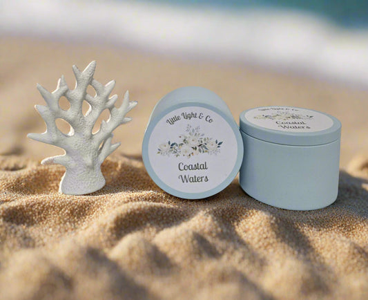 Two blue tins labeled 'Coastal Waters' by Little Lights & Co on a white surface with a white coral sculpture.