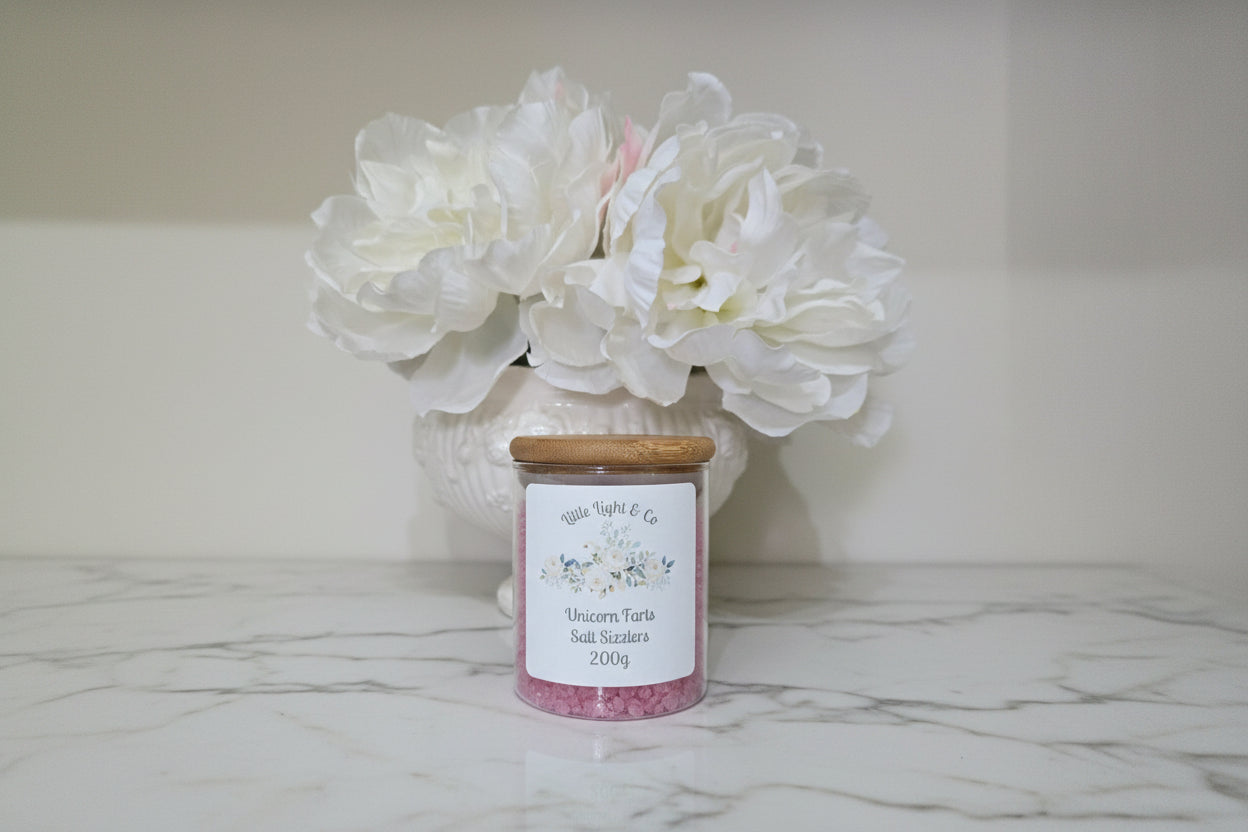 Jar of 'Unicorn Farts' salt with a wooden lid on a brown surface, flowers in the background.